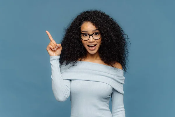 Förvånad lycklig kvinna med afro frisyr, pekar fore finger på tomt utrymme ovan, har öppnat munnen, bär glasögon och Jumper med nakna axlar, isolerade på blå väggen. Annons koncept — Stockfoto