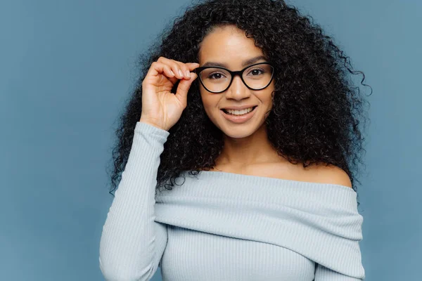 Headshot приємний дивлячись молодий афроамериканець Жінка тримає руку на рамі окуляри, має ніжний усмішкою, носить блакитний перемички, має мінімальний макіяж, здорова шкіра, ізольовані над синім фоном — стокове фото