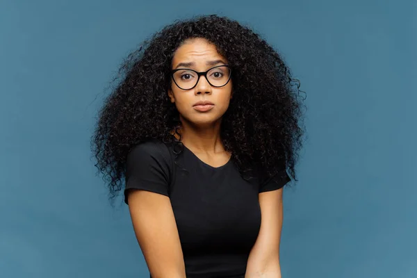 Foto av indignerade förbryllad afro amerikansk kvinna ser förvånansvärt vid kamera, bär casual svart t shirt och glasögon, poserar mot blå bakgrund. Människans ansiktsuttryck och känslor koncept — Stockfoto