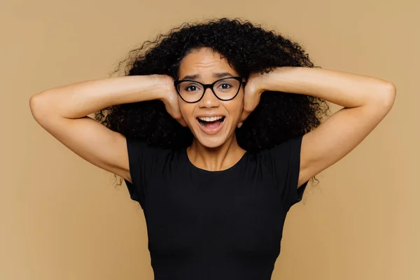 Jeune femme aux cheveux frisés afro couvre les oreilles, crie comme des demandes être calme, ne peut pas tenir le bruit, garde la bouche ouverte, porte des lunettes et t-shirt noir décontracté, isolé sur fond de studio brun . — Photo