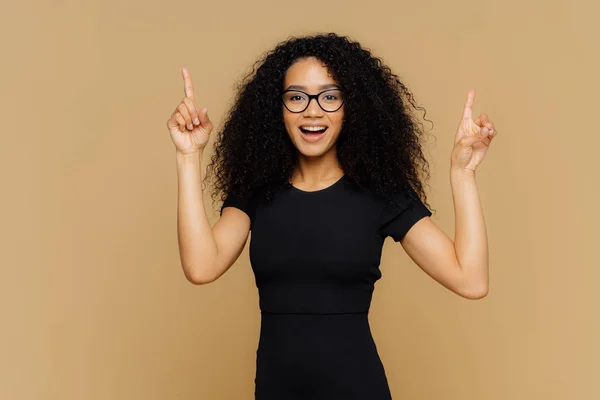 Regarde vers le haut. Heureuse femme afro-américaine pointe ci-dessus avec les deux index, a une silhouette mince, porte un t-shirt noir, promeut l'objet, isolé sur fond brun. Publicité et ethnicité — Photo