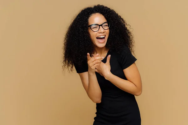 Внутрішній знімок переповненої жінки голосно сміється, зауважує смішну сцену, вказує на камеру, тримає рот широко розкритим, носить прозорі окуляри, чорну сорочку, ізольовані на коричневому тлі — стокове фото