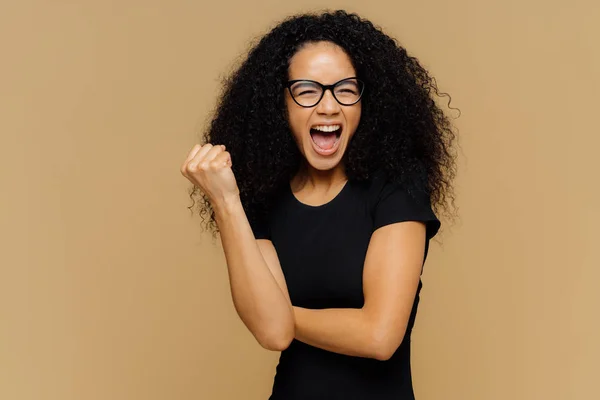 Animando mulher otimista aperta punho, gritos em voz alta, sendo muito emocional, apoia seu time de futebol favorito, veste camisa preta casual, fica sobre fundo marrom. Conceito de celebração — Fotografia de Stock