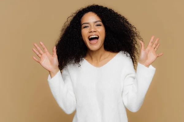 Emocional feliz afro-americano mulher com cabelo preto nítido, levanta as mãos, gestos ativamente, observa algo maravilhoso e impressionante, usa jumper branco macio, modelos sobre fundo marrom . — Fotografia de Stock