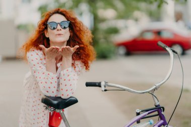Sevecen kızıl saçlı kadın sokakta sevgilisine hava öpücüğü gönderir, avuç içlerini ağzına yakın tutar, yuvarlak tonları giyer, elbise giyer, bisiklete yaslanır, açık havada aktif bir istirahat verir, sokakta poz verir. Eğlence zaman kavramı
