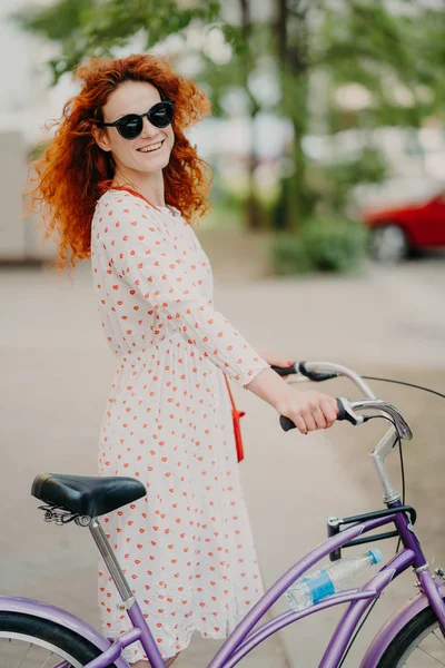 Smiling redhead woman in sunglasses and dress, rides bike through city during summer holidays, has rest or minute break, leads healthy lifestyle, enjoys recreation time, blurred urban background