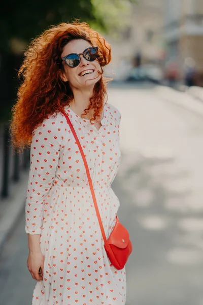 Optimistic foxy woman with glad expression, looks into distance, wears shades, long dress, strolls outdoor on street, enjoys nice summer day, has broad smile. People, lifestyle, rest and lifestyle