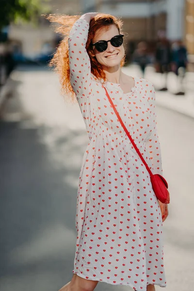 Mirthful young lady strolls on street, has long red hair, wears stylish dress, sunglasses, enjoys sunny day at weekend, poses over blurred street background, has nice mood. People and spare time