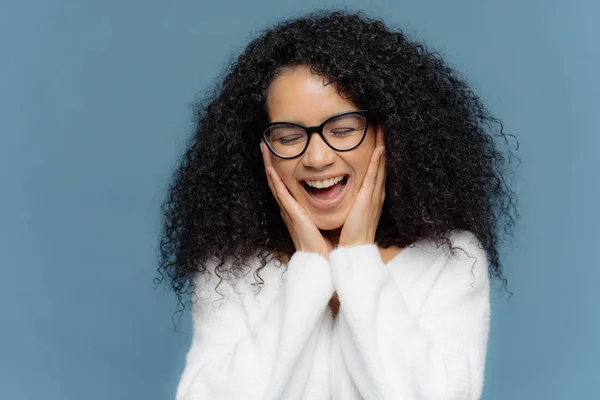 Overemotive female touches cheeks laughs from joy, keeps eyes closed, cannot stop giggling, has Afro hairstyle, dressed in white jumper, models against blue background. Ethnicity and emotions concept — Stock Photo, Image