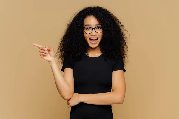 Brunette mixed race woman points aside with index finger, has glad curious expression, promots awesome product, dressed in casual clothes, isolated over brown background. People and advertisement — Stock Photo, Image