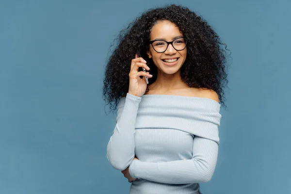 Indoor shot of positive lovely woman has telephone conversation, keeps mobile phone near ear, focused aside, wears casual jumper, isolated on blue background. People, technology, communication — Stock Photo, Image