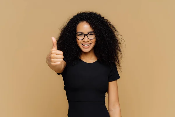 Waist up shot of satisfied supportive woman shows thumb up, cheers best friend, encourages for excellent efforts, wears glasses and black t shirt, stands against beige background. Body language