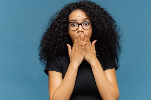 Photo of stupefied young female covers mouth, has bated breath, bugged eyes, Afro hair, cannot believe in shocking news, wears casual black t shirt, stands over blue background. Omg concept. — Stock Photo, Image