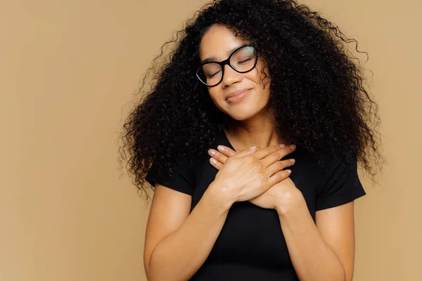 Touched lovely female with Afro hairstyle, keeps both palms on chest, has eyes shut from pleasure, hears heart piercing story, wears spectacles and casual t shirt, models against beige background — Stock Photo, Image