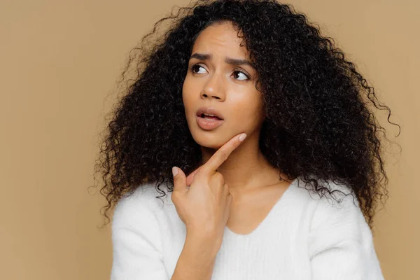 Modelo feminino intrigado mantém dedo indicador no queixo, olha cuidadosamente de lado, frustrou a expressão facial, cabelo afro encaracolado, usa jumper branco casual, isolado sobre fundo marrom. Fechar tiro — Fotografia de Stock