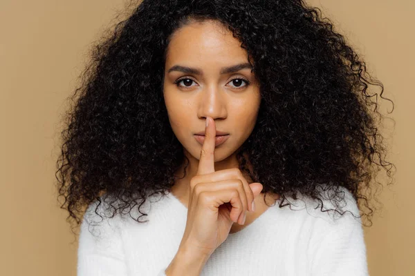 Imagen de mujer de piel oscura confiada seria hace gesto de calla, mantiene el dedo por encima de los labios, cuenta información secreta, pide no difundir rumores, vestido con traje casual, aislado sobre fondo beige — Foto de Stock