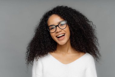 Carefree aşırı sevindi koyu tenli kadın mutlu gülüyor, yüksek ruh olmak, kafa Eğir, kıvırcık koyu saçlı vardır, beyaz Jumper giyer, komik bir şey duyuyor, gri stüdyo arka plan karşı modelleri.