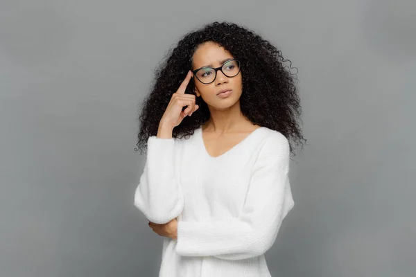 Troubled serious female keeps fore finger on temple, looks aside, has curly hair, being deep in thoughts, wears white jumper and optical glasses, isolated on grey background. People and thinking — Stock Photo, Image