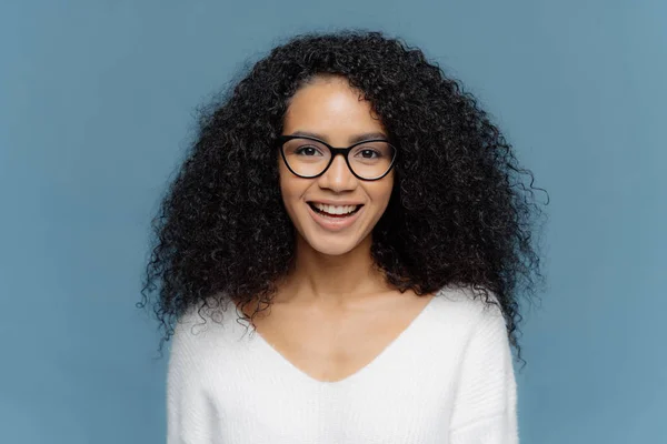 Photo de joyeuse femelle à la peau foncée aux cheveux touffus, souriant joyeusement à la caméra, se réjouit belle journée, vêtue d'un pull blanc, isolée sur fond bleu exprime positivité. Sentiments humains — Photo