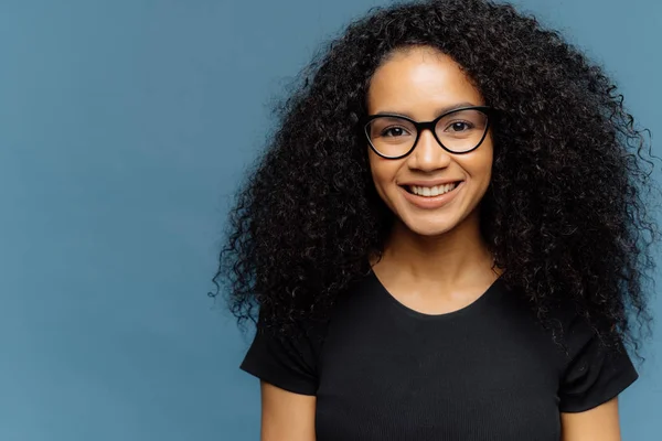 Headshot d'agréable femme à la peau foncée avec coupe de cheveux afro, porte des lunettes transparentes et t-shirt noir décontracté, exprime de bonnes émotions, se tient sur fond bleu, espace libre pour le slogan — Photo