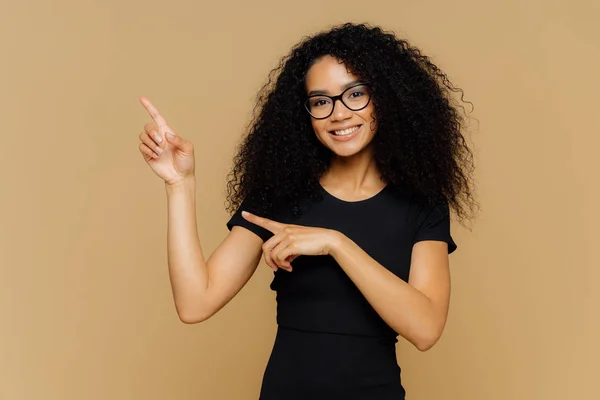 Photo isolée de femme afro-américaine heureuse pointe vers le haut et de côté, démontre l'espace de copie vierge pour votre publicité, porte des lunettes, t-shirt décontracté, a doux sourire sur le visage. Personnes et promo — Photo