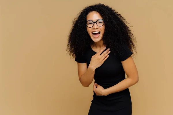 Magnifique mannequin à la peau foncée ne peut s'empêcher de rire, entend une histoire comique, garde la main sur le ventre, rit fort, vêtu de vêtements noirs, se dresse contre le mur du studio brun avec un espace vide pour l'information — Photo