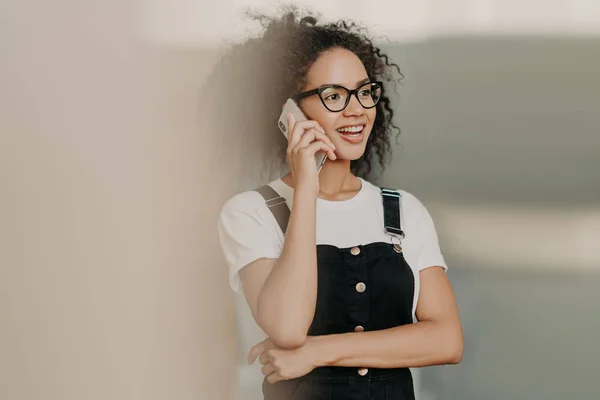 Smiling young curly woman with gentle smile, talks on mobile phone, has friendly conversation, wears casual white t shirt and overalls, focused aside. Leisure time, lifestyle, technology concept