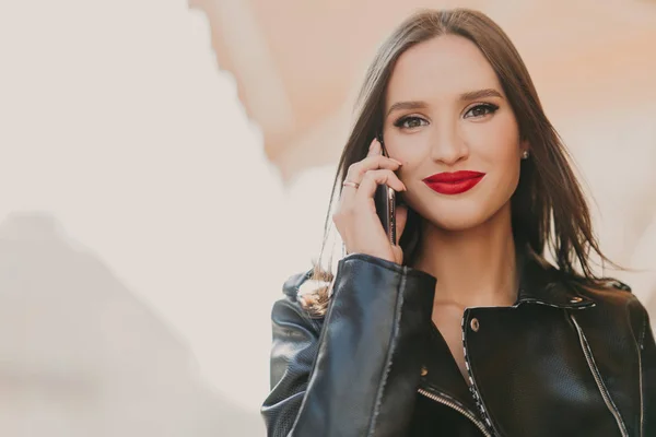 Attractive lovely female enjoys tariffs in roaming, talks on mobile phone with best friend, dressed in fashionable clothes, wears red lipstick, makeup, stands on blurred background of some building