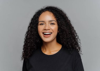 Mutlu koyu tenli Afro Amerikalı kadının yakın çekim olumlu gülüyor, iyi bir ruh hali içinde olmak, siyah rahat giysiler giymiş, gri arka plan üzerinde izole. İnsan duyguları ve hissetmek kavramı.