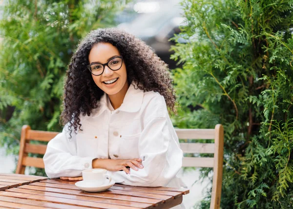 Relaxed cheerful Afro woman in white clothes, drinks coffee in outdoor cafeteria, sits at wooden table on chair against green thuja, has happy expression. People, lifestyle, spare time concept