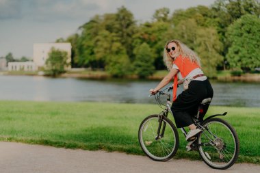 Neşeli bir kadının resmi, sıradan giyinmiş, bisiklete biniyor, yan tarafa bakıyor, mutlu bir ifade takınıyor, güneş gözlüğü takıyor, nehir kenarında poz veriyor, yeşil çimenler ve ağaçlar, arka planda bazı binalar. İnsanlar ve dinlenme
