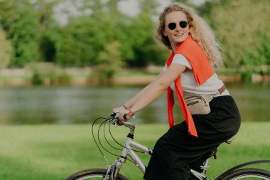 Aktif Kafkasyalı kıvırcık kadın yana bakıyor, spor bisikleti sürüyor, mesafeyi katediyor ve açık havada seyahat ediyor, günlük kıyafetler giyiyor, yeşil ağaçlı küçük bir nehrin yanında poz veriyor, sağlıklı bir yaşam tarzı var.