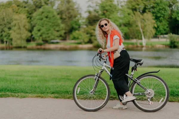 Aktif kadın bisikletçinin yatay görüntüsü tişört, siyah pantolon ve spor ayakkabı giyer, nehir kenarında durur, arka planda güzel manzaralı bisiklet pedallarının üzerinde ayaklarını tutar. Güneşli gün, aktif dinlenme