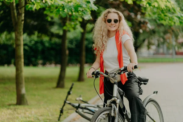 Aktif kıvırcık saçlı kadın bisikletçinin kendi aracıyla mesafeyi kat etmesine, güneş gözlüğü takmasına, günlük kıyafet giymesine yeşil ağaçlarla parkta poz vermesine, iyi duygular göstermesine sevindim. Bisiklet sürme kavramı