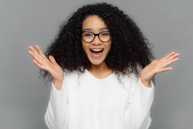 Samimi duygular ve duygular kavramı. İyimser güzel genç kadın elleri yayılır, hoş bir şey görür mutlu güler, kıvırcık gür saç modeli vardır, gözlük ve beyaz rahat jumper giyer