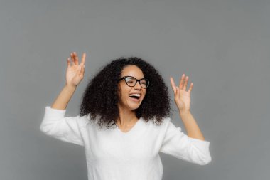 Overjoyed Afro Amerikalı kadın ellerini kaldırır, mutlu güler, hoş müzik sahiptir, optik gözlük ve beyaz rahat jumper giyer, gri arka plan üzerinde modeller, metin için yukarıdaki kopya alanı.