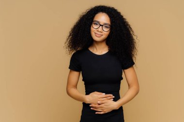 Kıvırcık saçlı Afro Amerikalı kadın memnun, göbek ellerini tutar, besleyici lezzetli akşam yemeği yedikten sonra iyi hissediyor, kamera mutlu görünüyor. Genç mutlu kadın hamilelik hakkında öğrenir.