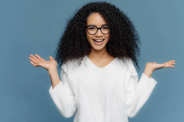 Jeune femme amicale positive à l'apparence afro, écarte les paumes, tient un objet invisible, a un sourire charmant, une peau saine, porte des lunettes et un pull blanc, isolé sur fond bleu . — Photo