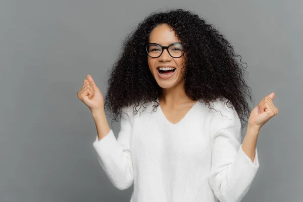 Femme afro-américaine ravie serre les poings avec triomphe, rit de bonheur, se réjouit du succès, porte des lunettes, pull blanc, acclamations pour l'équipe préférée, isolé sur fond gris — Photo