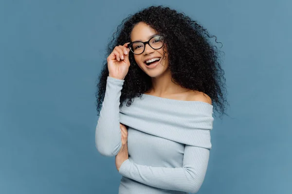 Isolerade skott av leende ung afrikansk amerikansk kvinna berör ram av glasögon, har glad uttryck, bär snygg tröja, isolerad på blå bakgrund, lycklig tillbringa ledig tid med vänner. Känslor — Stockfoto