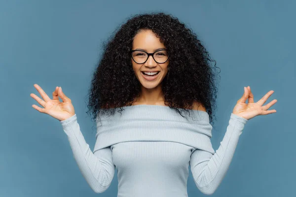 Positive African American lady makes okay gesture with both hands, meditates indoor, wears fashionable sweater and eyewear, being in good mood, isolated over blue background. Body language concept