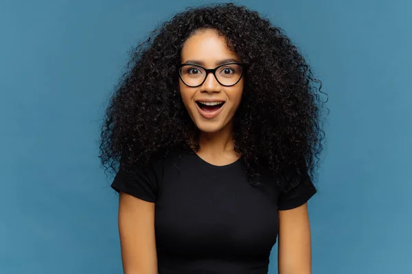 Agréablement surpris femme à la peau foncée garde la mâchoire baissée, regarde avec intérêt, a les cheveux bouclés, porte un t-shirt noir, des modèles contre le mur bleu. Afro American dame regarde avec choc, a soufflé souffle — Photo