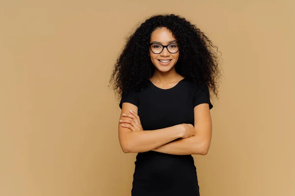 Slim nöjd kvinna med afro frisyr, bär svart casual kläder, optiska Glasögon, har förtroende uttryck, lyssnar samtalspartner, isolerade på beige bakgrund. Söt tonårs flicka njuter av livet. — Stockfoto