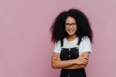 Afro saçlı, kollarını katlamış, gözlük takmış, beyaz tişört ve siyah tulum giymiş, kameraya sevinçle bakan, metin için mor arkaplanda duran güzel bir genç kızın fotoğrafı.