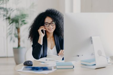 Akıllı telefondan arayan Afro-Amerikan kadınlarının fotoğrafı not defterine not alıyor, çalışma sürecinde yer alıyor, bilgisayarla masaya oturuyor, gözlük takıyor, resmi kıyafetler giyiyor, telefon iletişimi kuruyor.
