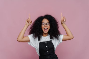 Mutlu Afro-Amerikan kadınlarının fotoğrafı her iki işaret parmağını da yukarıda gösteriyor, muhteşem fotokopi alanını gösteriyor, optik gözlük takıyor ve moda kıyafetler giyiyor, menekşe arkaplan üzerinde poz veriyor. Yukarıya bak.