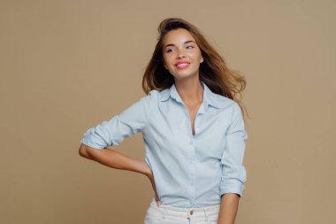 Shot of pretty cheerful European woman keeps one hand on waist, looks aside, has charming smile wears shirt and trousers, has slim perfect figure, enjoys shopping day, isolated over brown background clipart