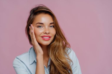 Hoş görünümlü kadın modelin fotoğrafı avuç içi ile dokunur, yumuşak dişli gülümsemesi vardır, makyaj yapar, günlük gömlek giyer, mor duvara yaslanır, reklamınız için alan kopyalarsınız.