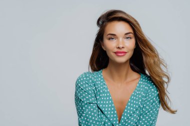 Pozitif genç kadının uzun dalgalı saçları, makyajı var, turkuaz polkadot bluz giyiyor, kameraya düzgün bakıyor, beyaz arka plana karşı modeller, reklamınız için kopyalama alanı.