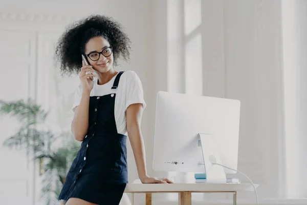 Pleasant looking African American female has telephone conversation, asks for advice before job interview, dressed in stylish clothes, poses near workplace with modern technology. Communication